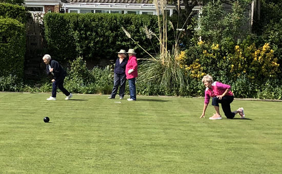 Four ladies on the green. In the foreground one has just delivered a bowl which can be seen travelling to the left of the picture. In the background, two ladies stand watching while a third bowls